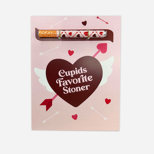 A romantic and whimsical greeting card in pastel pink, adorned with a heart-shaped chocolate box reading 'Cupid's Favorite Stoner,' accented with playful arrows, hearts, and a joint, combining elements of love and cannabis culture