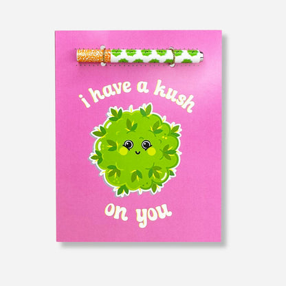 A cute and endearing greeting card with a soft pink background, adorned with a smiling cartoon cannabis bud and the pun 'I have a kush on you,'.