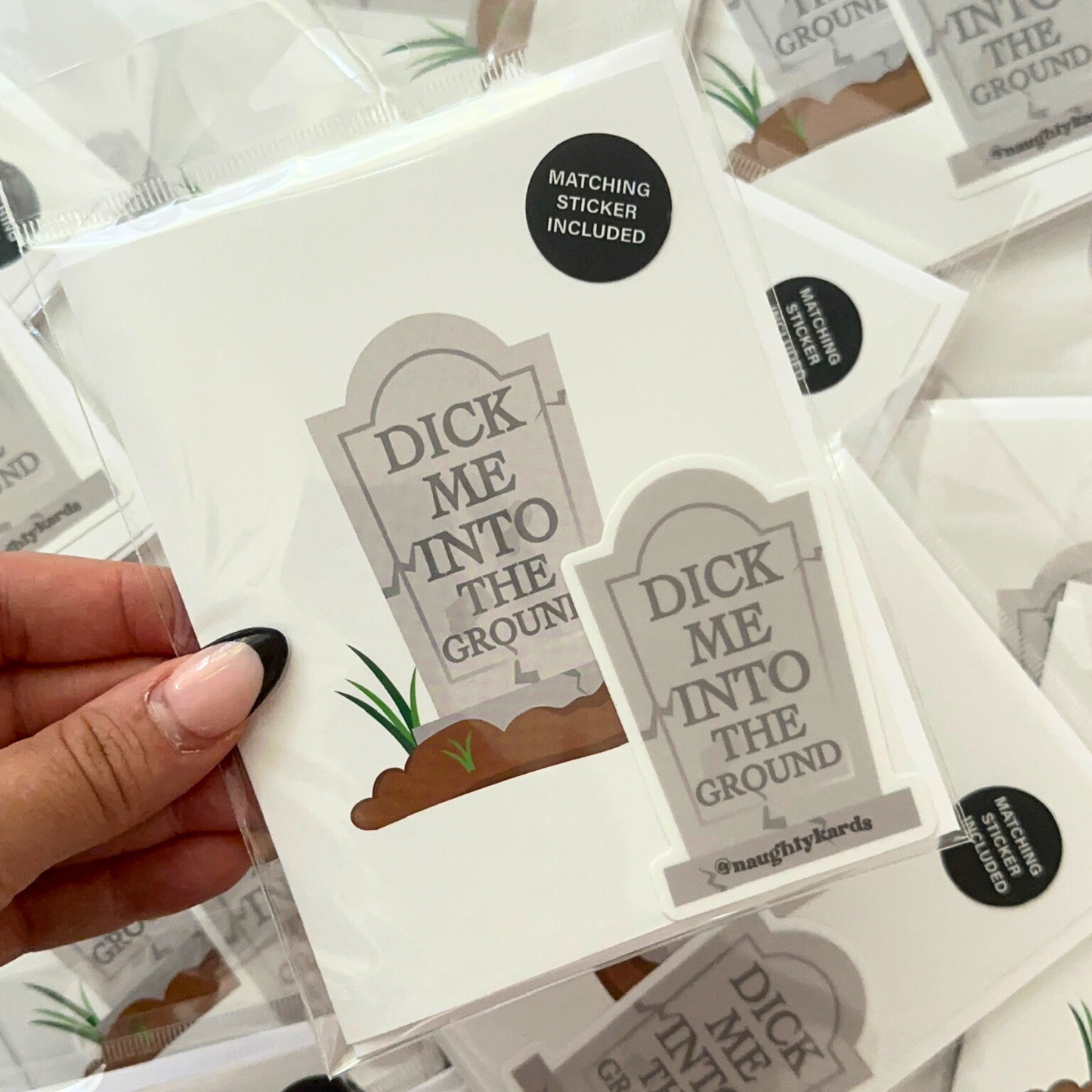 Hand holding a 'Dick Me Into the Ground' greeting card set, featuring a tombstone design with matching sticker included in packaging, part of the NaughtyKards collection.