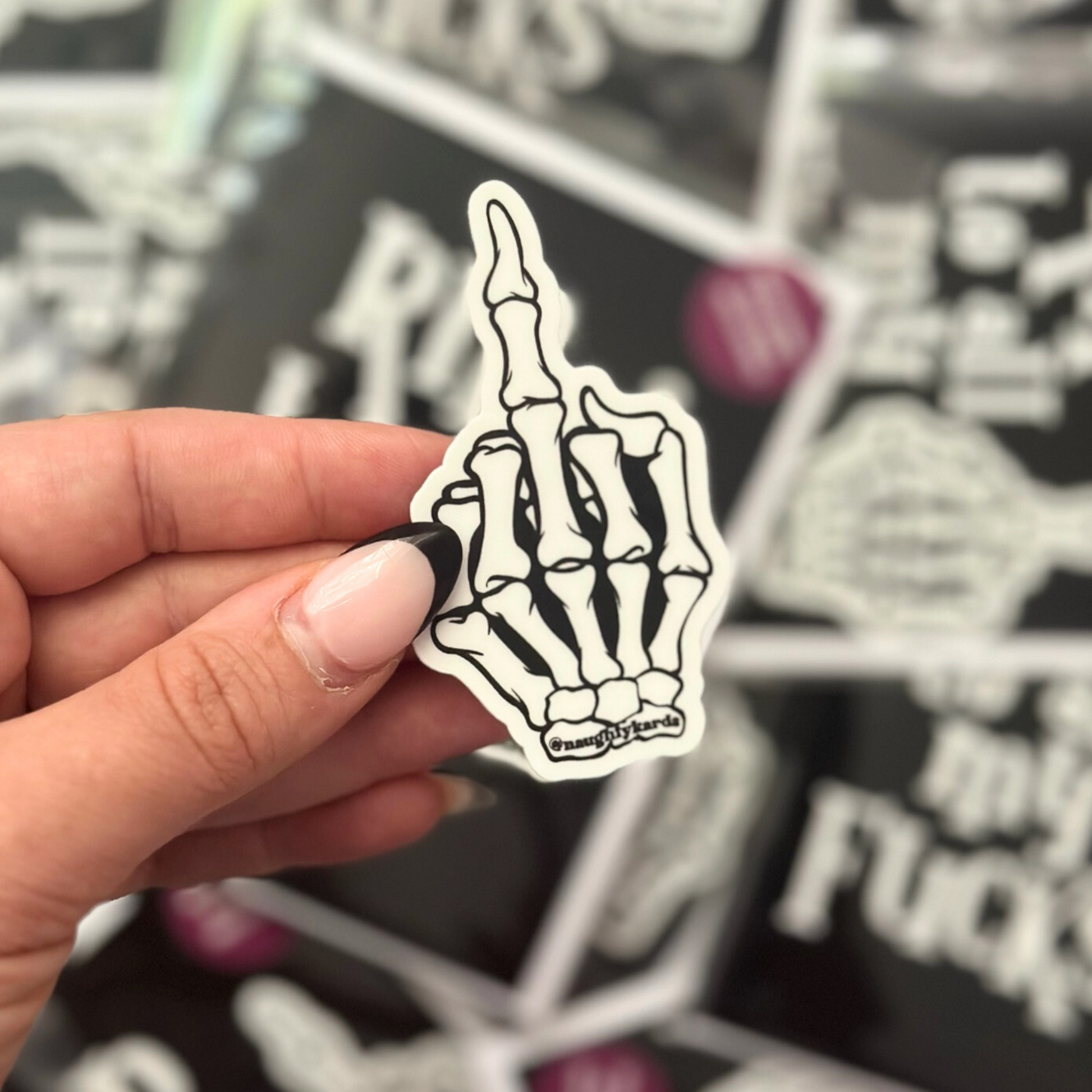 Close-up of a hand holding a small sticker featuring a skeleton hand with the middle finger raised, with other similar stickers displayed in the background.