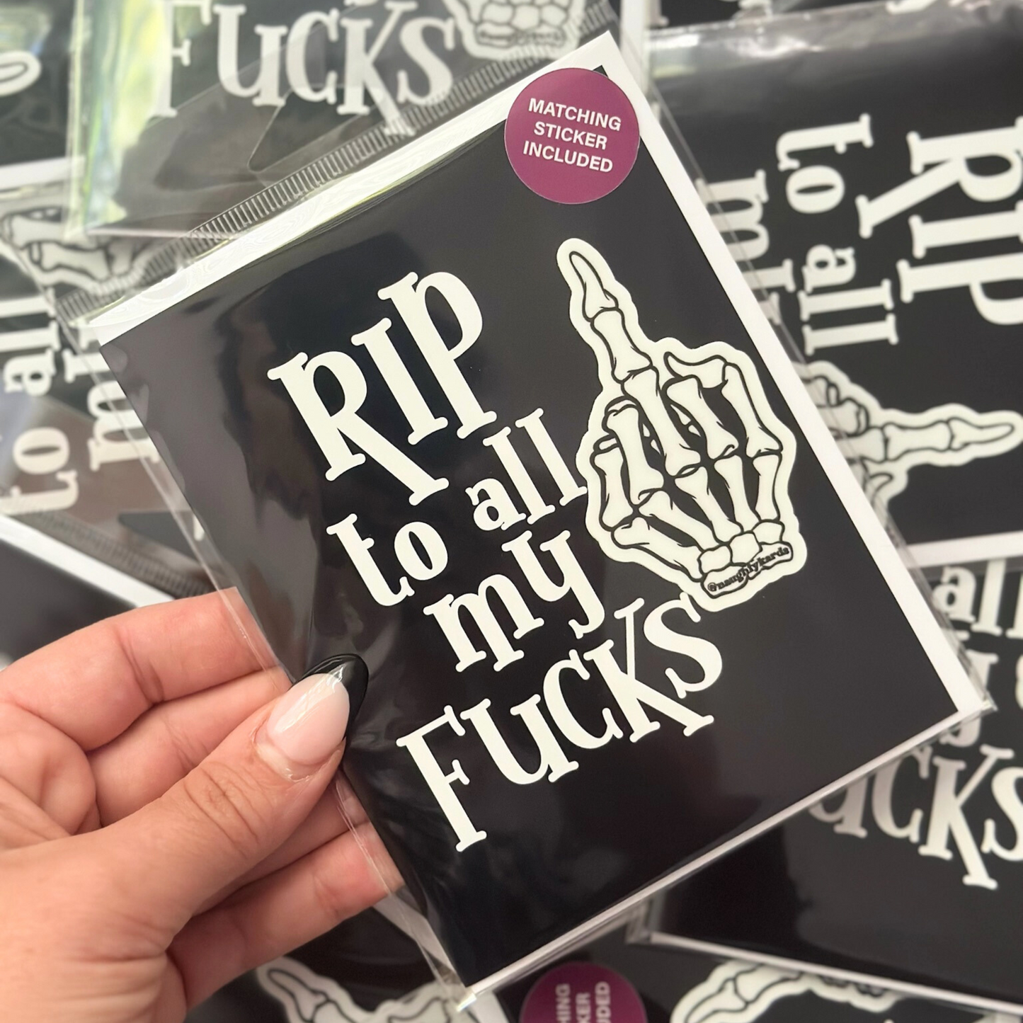 Close-up of a hand holding a greeting card with the words "RIP to all my fucks" in bold white letters. The card includes a matching sticker featuring a skeleton hand with the middle finger raised.