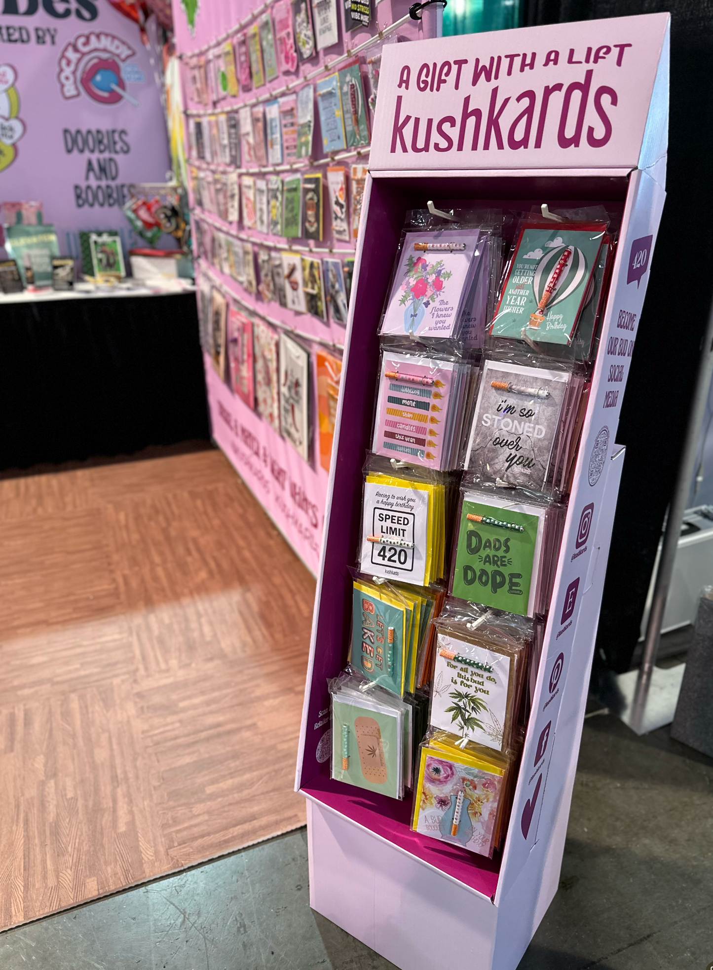 KushKards cardboard display at a trade show, filled with colorful greeting cards, with the brand’s signage 'A GIFT WITH A LIFT' visible on top, set against an exhibit backdrop featuring similar products.