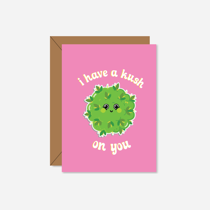 A tiny pink Valentine's Day card featuring a cute, smiling green bud illustration with the phrase "I Have a Kush On U" in playful white script, accompanied by a kraft paper envelope.